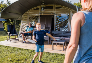 Dreng spiller badminton foran en safaritelt, mens en voksen og endnu en person slapper af på terrassen.