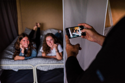 Twee jonge dames poseren lachend op een bed in een safaritent terwijl iemand een foto van hen maakt met een smartphone.