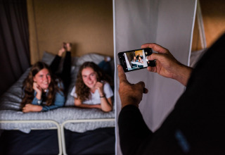 Twee jonge dames poseren lachend op een bed in een safaritent terwijl iemand een foto van hen maakt met een smartphone.