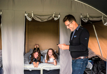 Un homme sourit avec un téléphone devant deux filles qui rient sur un lit dans une tente safari.