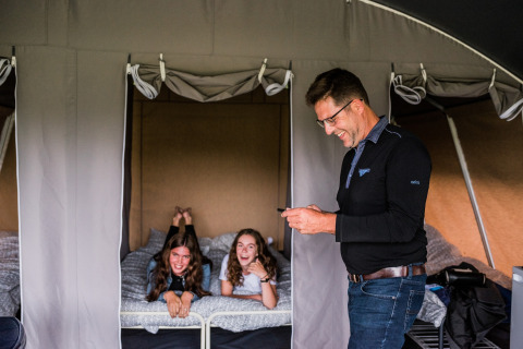 Un homme sourit avec un téléphone devant deux filles qui rient sur un lit dans une tente safari.