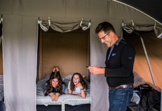 Een man glimlacht met een telefoon terwijl twee meisjes lachen op een bed in een safaritent.