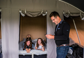 Un homme sourit avec un téléphone devant deux filles qui rient sur un lit dans une tente safari.