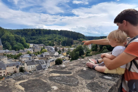 Un homme avec un enfant sur un mur observe une vue de village entouré de forêts et de collines verdoyantes.