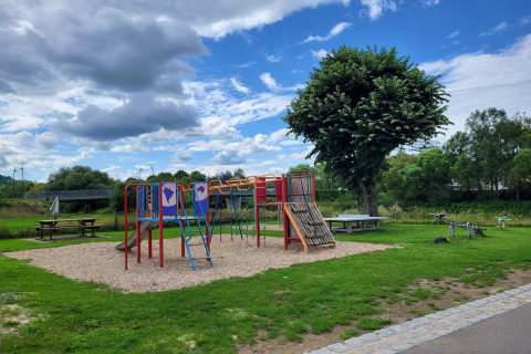 Foto van een speeltuin met glijbaan, klimtoestel en picknicktafels, omgeven door gras en bomen.