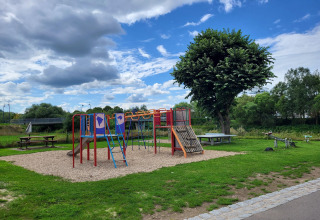 Foto van een speeltuin met glijbaan, klimtoestel en picknicktafels, omgeven door gras en bomen.