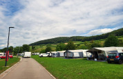 Photo d’un camping avec tentes safari, voitures et collines vertes à l’arrière-plan sous un ciel nuageux.