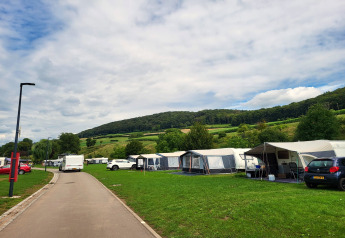 Photo d’un camping avec tentes safari, voitures et collines vertes à l’arrière-plan sous un ciel nuageux.