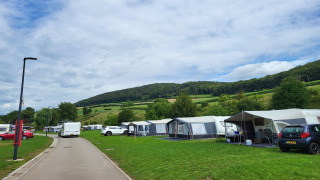 Foto van een camping met safaritenten, auto's en groene heuvels onder een bewolkte lucht.