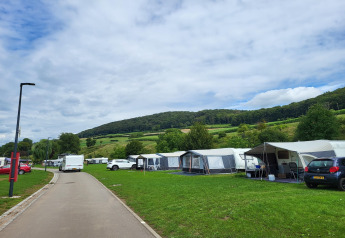 Foto van een camping met safaritenten, auto's en groene heuvels onder een bewolkte lucht.
