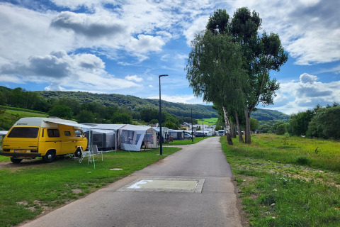 Een moderne camping met safaritenten en een gele camper, omgeven door groene heuvels en hoge bomen.