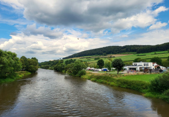 Vue depuis une tente safari sur une rivière, des champs verdoyants, des collines et des bâtiments sous les nuages.