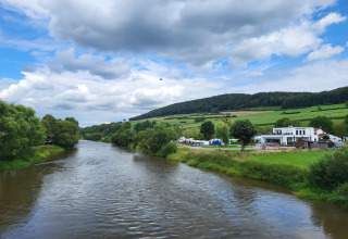 Uitzicht vanaf een safaritentterrein op een rivier, groene velden, heuvels en gebouwen onder de wolken.