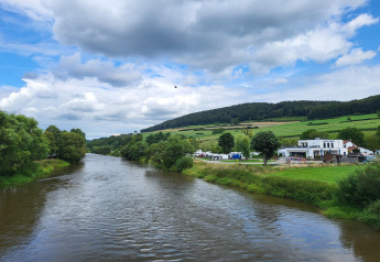 Uitzicht vanaf een safaritentterrein op een rivier, groene velden, heuvels en gebouwen onder de wolken.