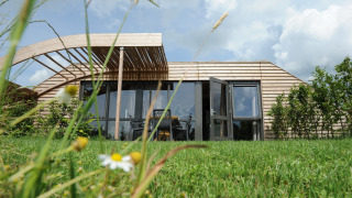 Et moderne træhus med store vinduer, græsplæne og vilde blomster i forgrunden under en blå himmel.
