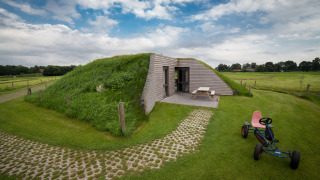 Græsbeklædt lodge med træfacade og picnicbord ved Koe in de Kost 4, omgivet af grønne marker.