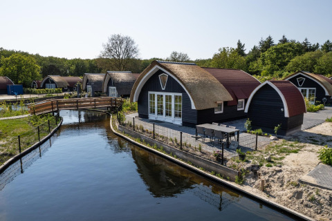 Thatched lodges at 't Landgoed Baars 1 by a canal, surrounded by lush trees and natural scenery.