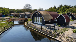 Cabañas con techo de paja en 't Landgoed Baars 1 junto a un canal, rodeadas de vegetación y naturaleza.