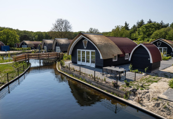 Thatched lodges at 't Landgoed Baars 1 by a canal, surrounded by lush trees and natural scenery.