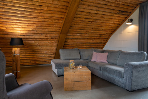 Hyggelig stue med skråt træloft, grå sofa og rustikke detaljer i Koe in de Kost 2 lodge i Holland.