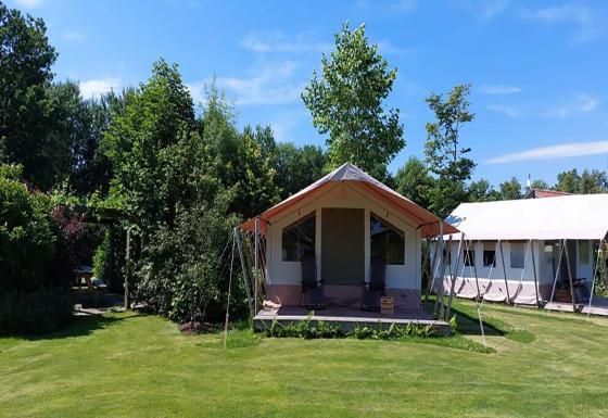 Safari tent 'Noflik' set in a lush green landscape with trees and grassy lawn beneath a clear blue sky.