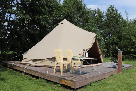 Glamping-Unterkunft mit einem großen Zelt auf Holzplattform, zwei Stühlen und Tisch in grüner Umgebung.
