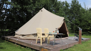 Glamping-oplevelse med et stort telt på en træplatform, to stole og et bord, omgivet af grøn natur.