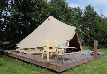 Glamping-oplevelse med et stort telt på en træplatform, to stole og et bord, omgivet af grøn natur.