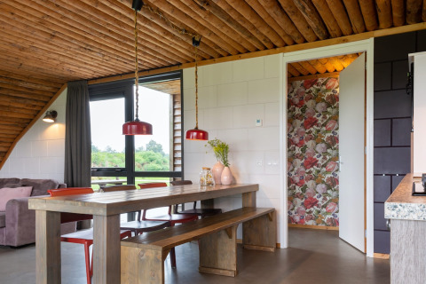 Zona pranzo in un lodge con soffitto in legno, sedute a panca, lampade rosse e carta da parati floreale.