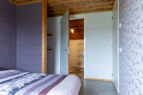 Camera da letto con pareti viola, carta da parati floreale, travi in legno e porta aperta sul bagno.