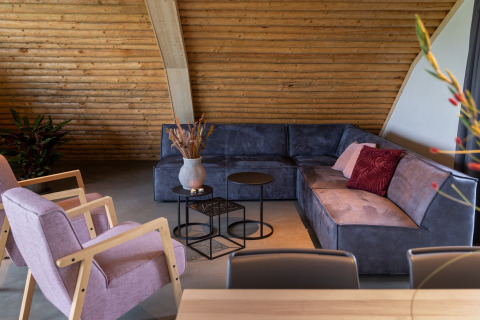 Woonkamer in Koe in de Kost 4, Nederland, met blauwe hoekbank, paarse stoelen en houten wandbekleding.