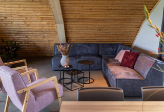 Woonkamer in Koe in de Kost 4, Nederland, met blauwe hoekbank, paarse stoelen en houten wandbekleding.