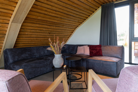 Woonkamer in lodge Koe in de Kost 4 in Nederland, met hoekbank, kussens, vaas en groot raam.