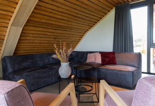Woonkamer in lodge Koe in de Kost 4 in Nederland, met hoekbank, kussens, vaas en groot raam.