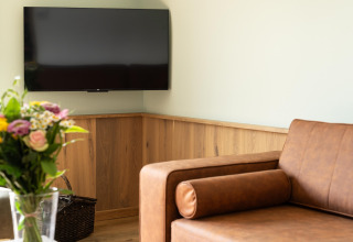 Modern lounge with brown sofa, fresh flowers, and wall-mounted TV at 't Landgoed Baars 1 in the Netherlands.