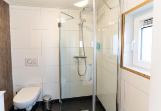 Modern bathroom with glass shower, wall-mounted toilet and window at ‘t Landgoed Baars 1 lodge in the Netherlands.