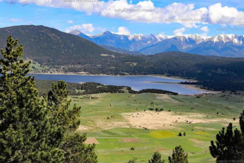 View of a lake surrounded by green fields, forests, and distant snowy mountains under a blue sky.