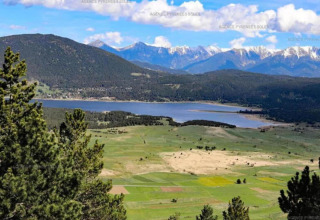 View of a lake surrounded by green fields, forests, and distant snowy mountains under a blue sky.