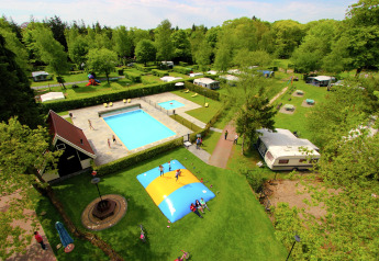 Luftaufnahme eines Glampingplatzes mit Pool, Hüpfkissen, Wohnwagen und viel Grün in idyllischer Natur.