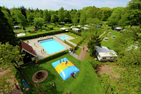 Luchtfoto van een glamping met zwembad, springkussen, caravans en groene zones omringd door bomen.