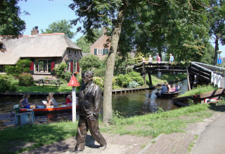 Gezellige lodge aan een kanaal met bootjes, brug, standbeeld en bezoekers op een zonnige dag.