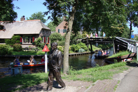 Sfeervolle lodge aan een kanaal met bootjes, brug, standbeeld en bezoekers op een zonnige dag.