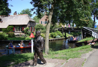 Sfeervolle lodge aan een kanaal met bootjes, brug, standbeeld en bezoekers op een zonnige dag.