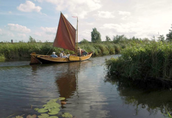 Eine Gruppe Menschen segelt in einem traditionellen Holzboot mit rotem Segel durch einen ruhigen Kanal.