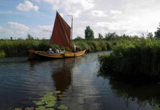 Een groep mensen vaart in een traditionele houten boot met rood zeil op een rustige, met riet omzoomde vaart.
