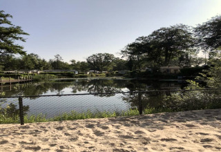 Uitzicht op een sereen meer en bomen vanop een zandplek met hek bij een glampingverblijf.