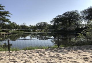 Uitzicht op een rustig meer en bomen vanaf een zandstrand met hek bij een glamping accommodatie.