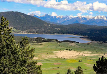 Panoramablick auf grüne Felder, einen klaren See und schneebedeckte Berge unter blauem Himmel.
