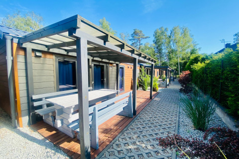 Udendørs foto af moderne glamping hytte med overdækket terrasse, bænk og havegang under blå himmel.