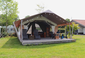 Safari tent with a wooden deck and outdoor furniture, set on a grassy lawn surrounded by trees.